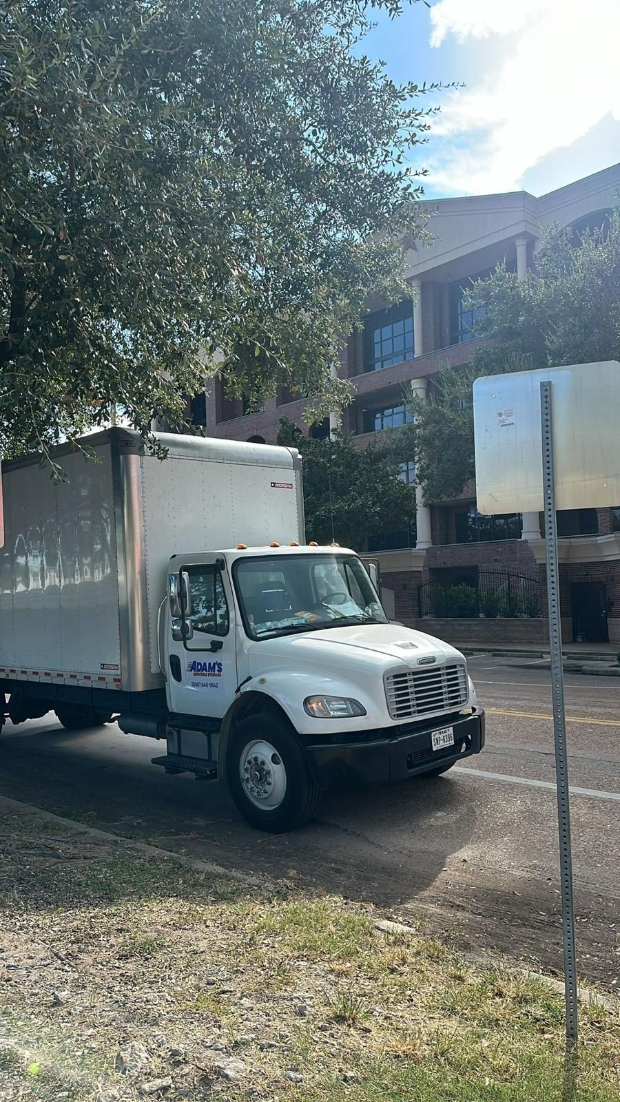 White delivery truck parked on a street with a brick building and trees in the background.